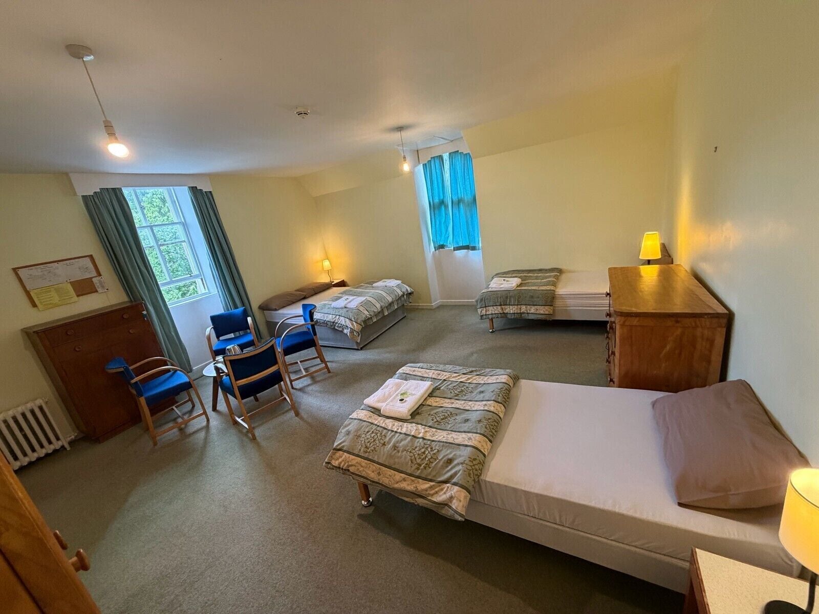 A hotel room with a bed, two smaller beds, a desk, a chair, a radiator, and a window with curtains.