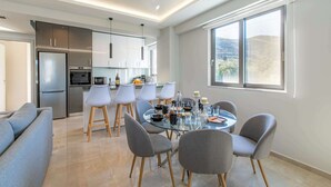 Une cuisine moderne dotée d’appareils électroménagers en acier inoxydable, un coin repas avec une table en verre et des chaises grises, et une grande fenêtre offrant une vue sur l’extérieur.