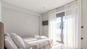 Une chambre moderne avec un grand lit, un téléviseur à écran plat fixé au mur et une porte-fenêtre coulissante donnant sur un balcon.
