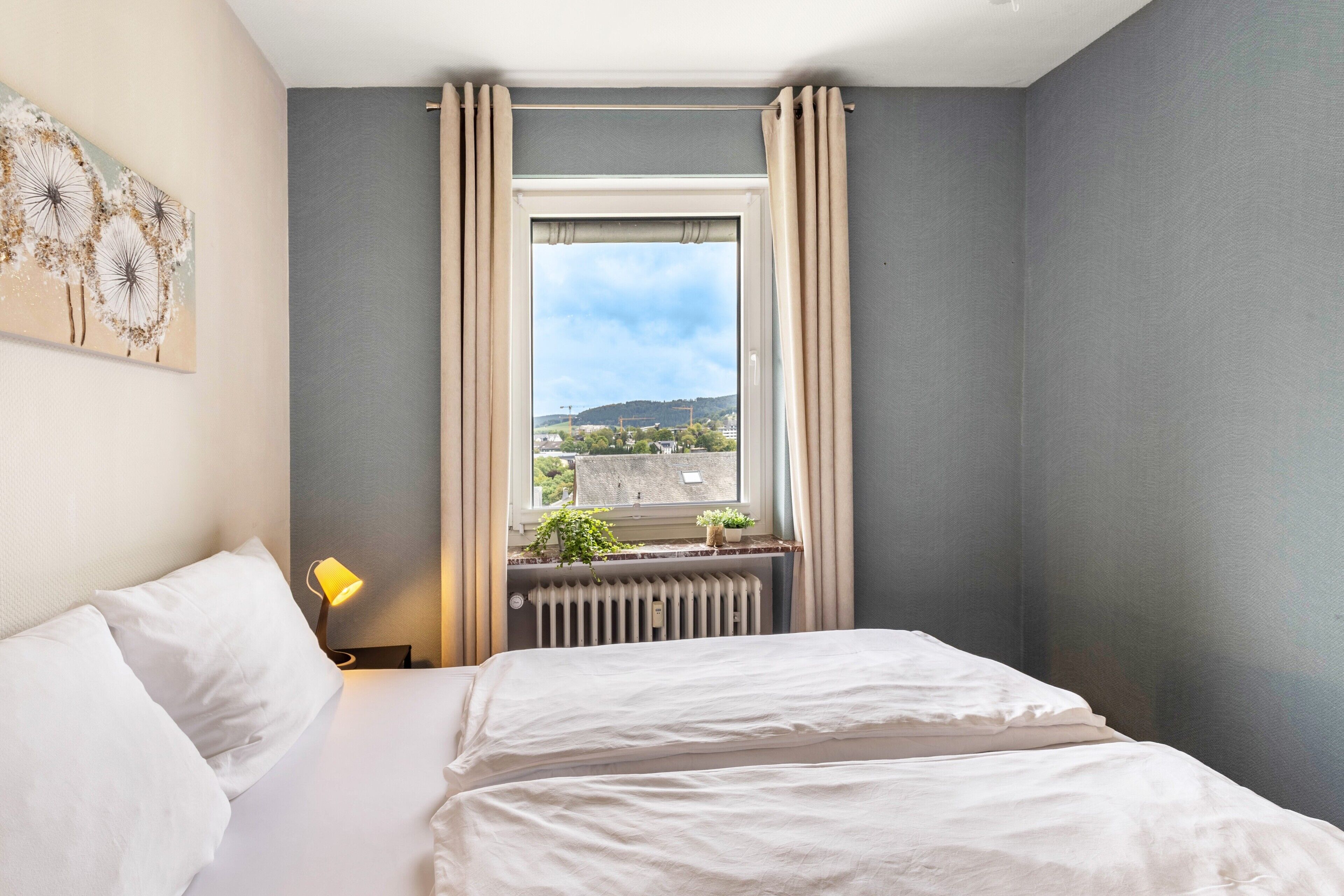 Une chambre d’hôtel avec un lit, une fenêtre avec des rideaux, un radiateur et une lampe fixée au mur.
