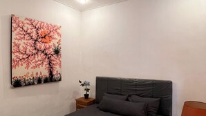 A modern bedroom with a grey bed, a nightstand with a lamp, and a floral painting on the wall. - Ajik Guest House (Ubud)