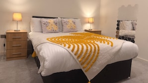 A neatly made bed with a patterned blanket, two bedside tables with lamps, a wooden nightstand, and a wall-mounted towel rack. - New Apartment - Sleeps 4 - Parking - City Centre (Bradford)