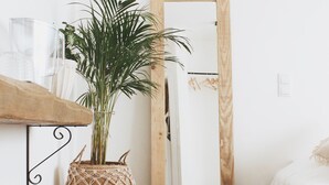 A room with a wooden floor, a bed, a small stool, a potted plant, a wall-mounted lamp, and a wooden door.