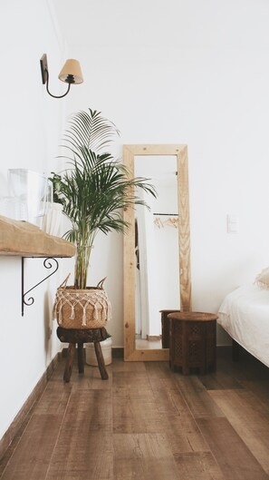 Une pièce avec un plancher en bois, un lit, un petit tabouret, une plante en pot, une lampe fixée au mur et une porte en bois.