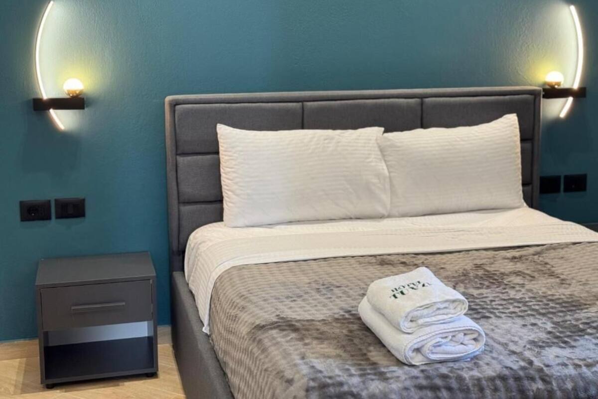 A neatly made bed with a grey headboard and white pillows, a small bedside table, and a folded towel on the bed.