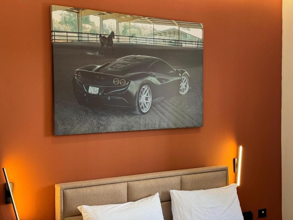 Un dormitorio con un cuadro grande enmarcados de un coche deportivo en la pared, una cama con ropa de cama blanca y una mesita de noche con una lámpara.