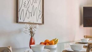 Ein Esstisch mit weißen Schalen und Früchten, eine Vase mit Trockenblumen und ein gerahmtes Schild an der Wand.