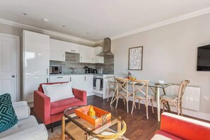 A modern kitchen with white cabinets, a dining area with a glass table and wooden chairs, and a living room with a red sofa and a glass coffee table. - 2BR Apt Near Windsor Castle Free Park By Tempstay (Windsor)