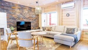 A modern living room with a stone wall, a sofa, a TV, and a dining area with chairs and a table. - Villa Pyry (Sotkamo)