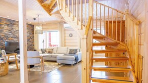A spacious living room with a wooden staircase, stone wall, and a large TV. - Villa Pyry (Sotkamo)