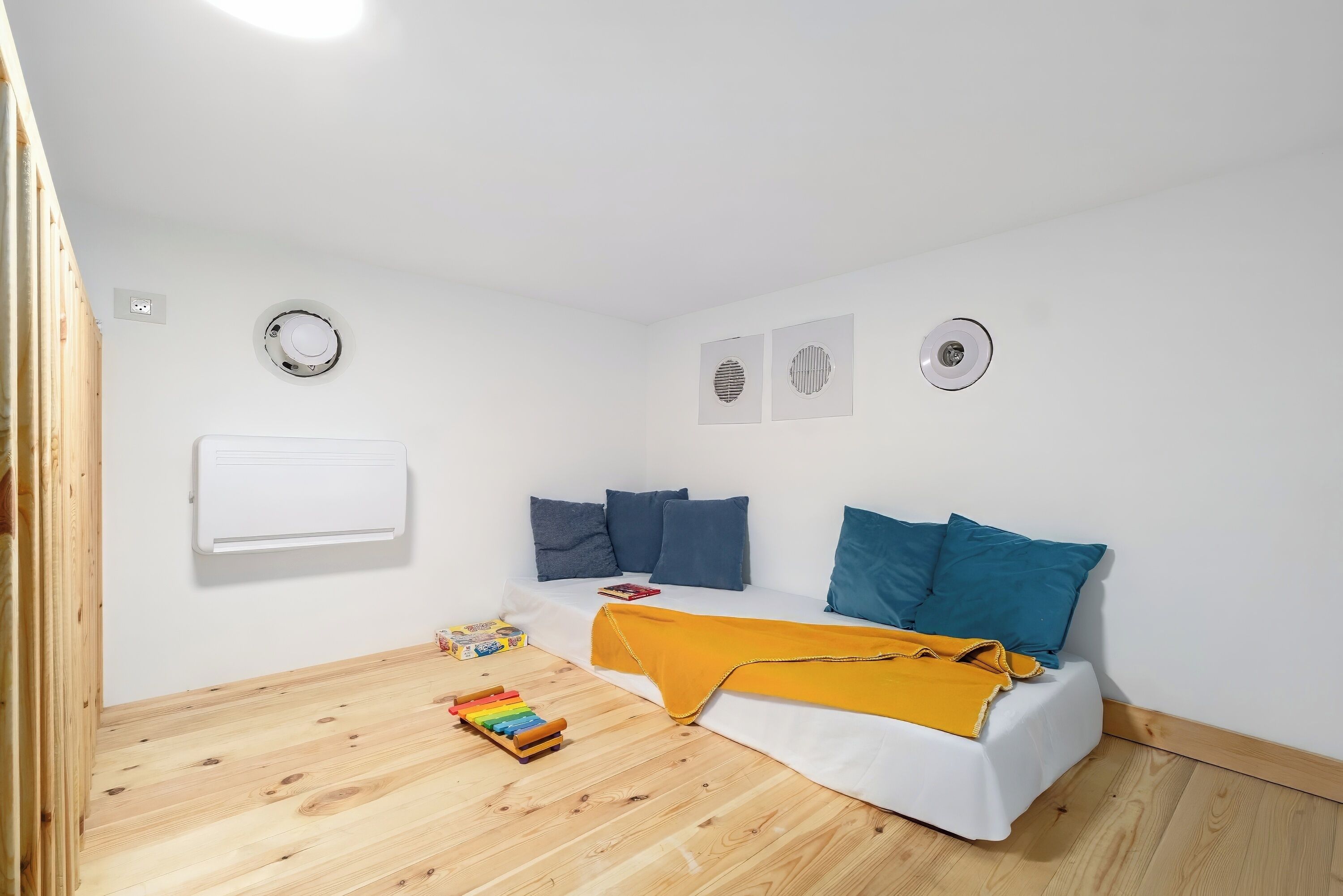 A small, bright room with a bed, a white air conditioner, and a wooden floor.