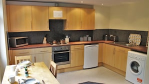 A compact kitchen with wooden cabinets, a microwave, oven, dishwasher, and washing machine. - Student Only Queens Court (Leicester)