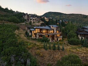 Una casa moderna de varios pisos con amplios ventanales y fachada de piedra, rodeada de árboles y colinas.