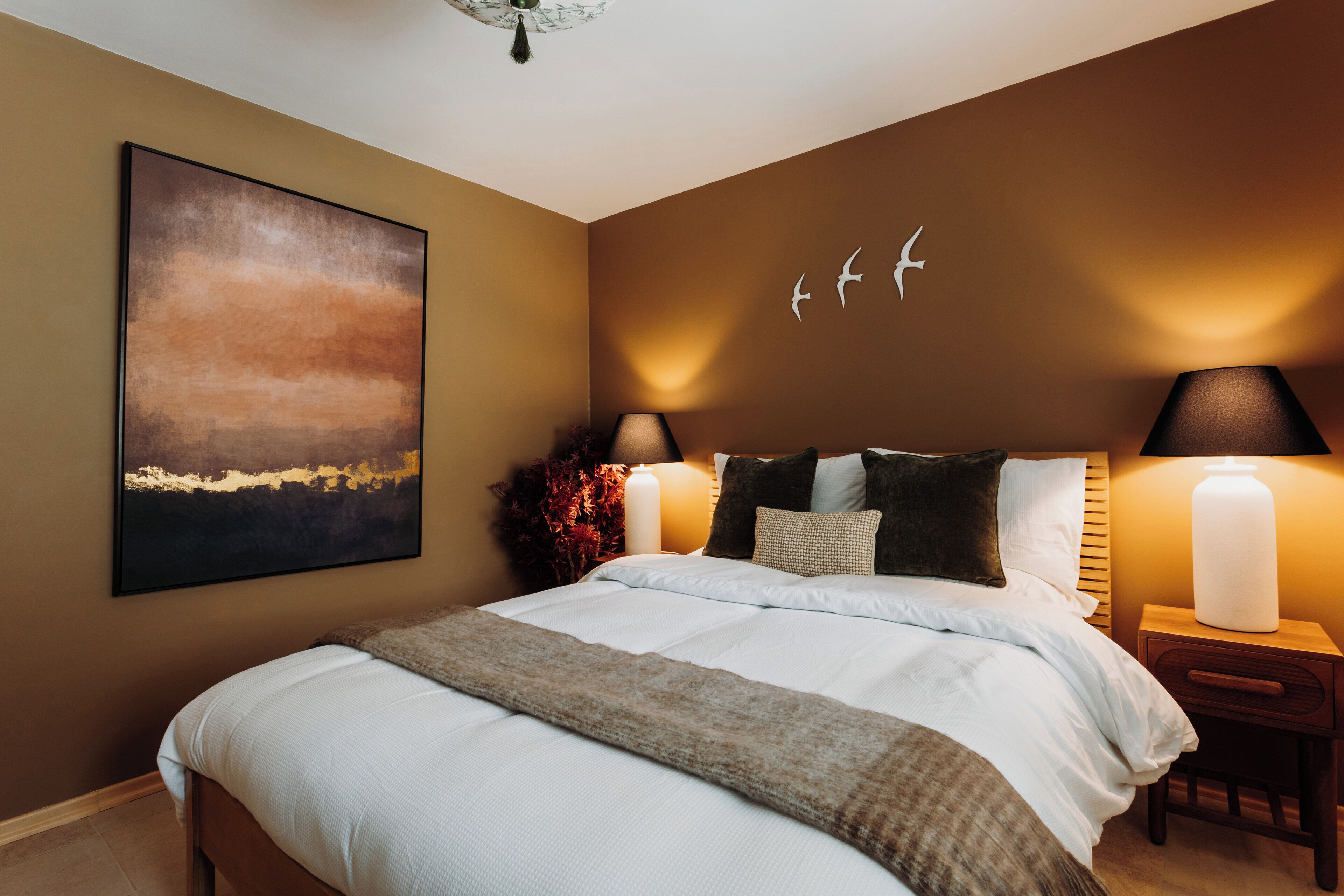 A bedroom with a large bed, two bedside tables with lamps, a wall-mounted artwork, and a decorative wall piece.