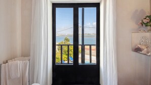 Quarto com vista para um corpo d'água e uma ponte, porta preta, cortinas brancas, porta-toalhas e prateleira com objetos decorativos.
