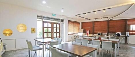 Un comedor amplio con varias mesas y sillas, ventanas grandes con persianas, y una zona de cocina al fondo.