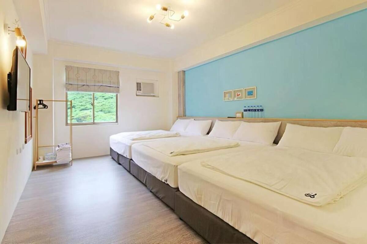A hotel room with two beds, a window with a view of greenery, a bedside table, and a wall-mounted TV.