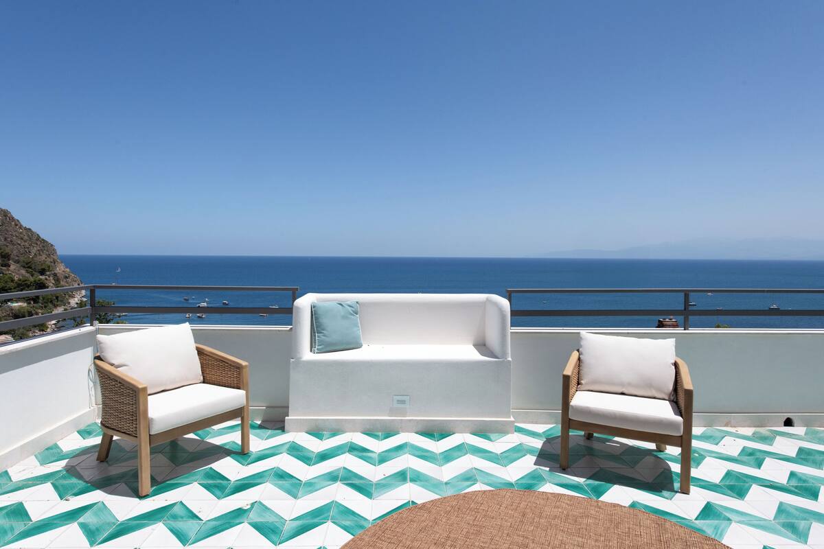 A terrace with two wicker chairs and a white sofa, a patterned floor, and a view of the sea.
