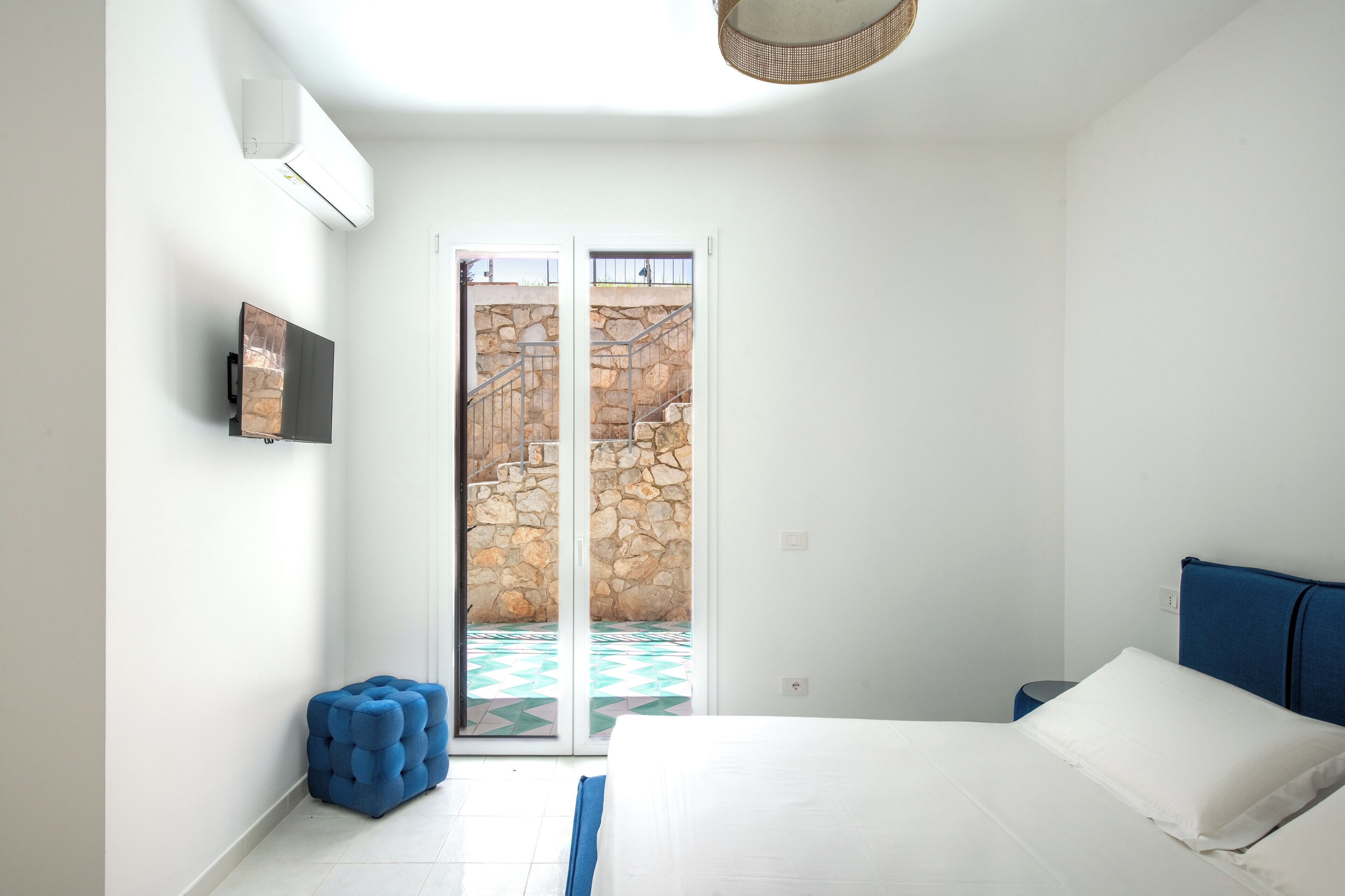 Una camera da letto con un letto, una televisione, un poggiapiedi blu e un muro in pietra visibile attraverso le porte scorrevoli.