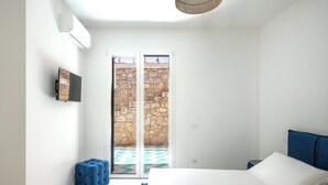 A bedroom with a bed, a TV, a blue ottoman, and a stone wall visible through the sliding doors. - Sea Frame Design Apartments with Pool by Wonderful Italy (Santa Flavia)