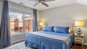 A bedroom with a large bed, two bedside lamps, a ceiling fan, and a view of buildings through a window. - Downtown Condo - 2BR, 2BA, Walk to Everything! (Raleigh)