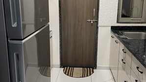 Eine Küche mit einem Zwei-Türen-Kühlschrank, einer Holztür, weißen Fliesenwänden und einem Fenster mit Spiegel sowie einem gefliesten Boden.