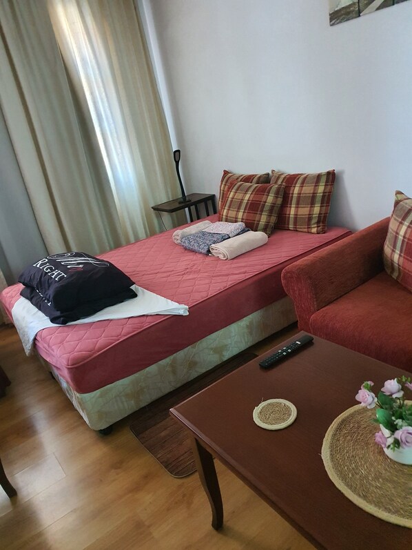 Una habitación con una cama, un sofá rojo, una mesita con un control remoto y una ventana con cortinas.