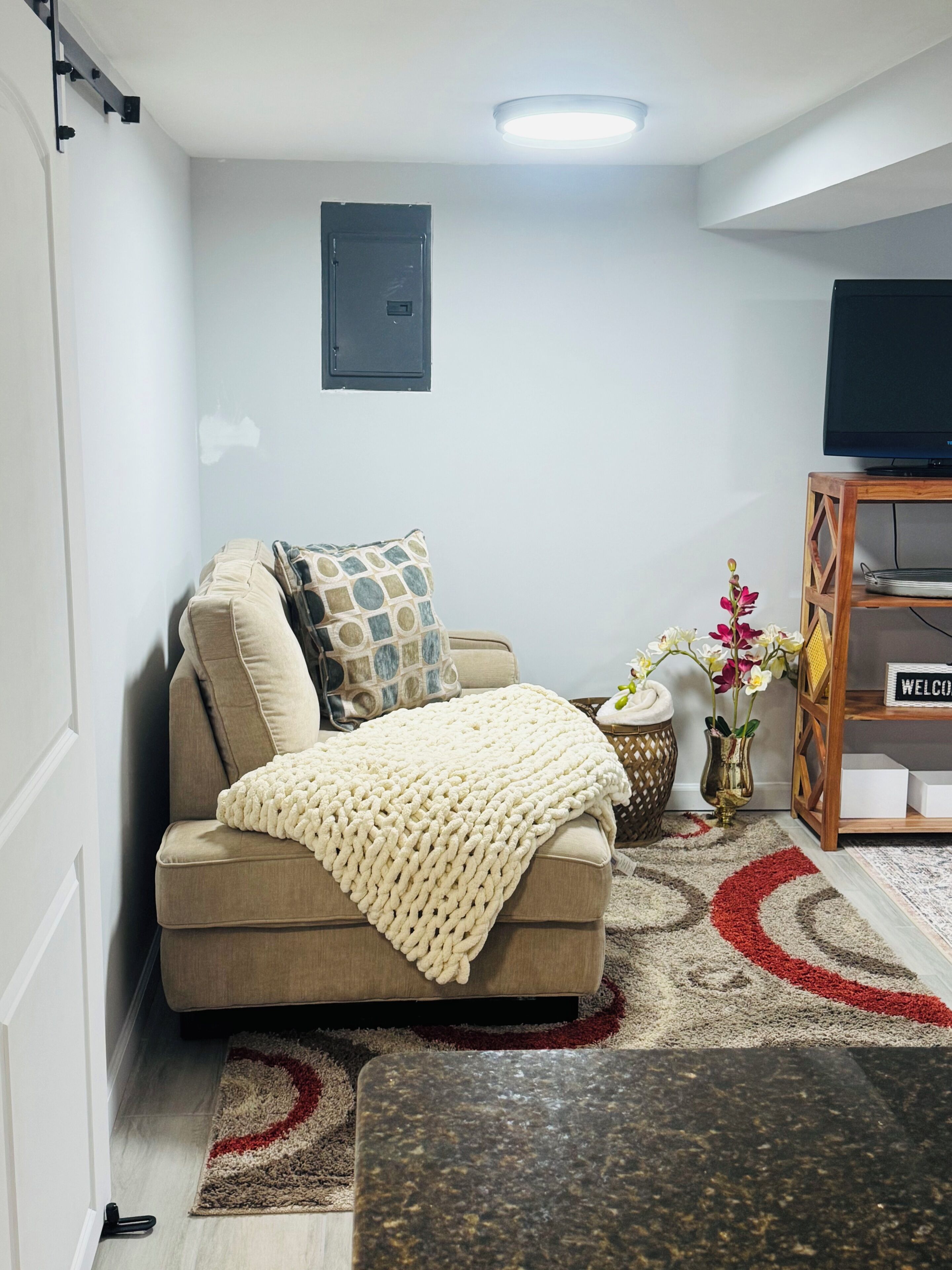Una sala de estar con un sofá color beige, cojines estampados, una manta tejida, una mesita de noche con un jarrón de flores, un aparador de madera con un cartel que dice 'WELCOME' y un televisor sobre un mueble de madera.