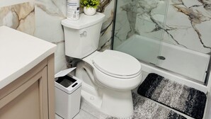Un baño con inodoro, ducha con mampara de vidrio, azulejos de mármol, una planta pequeña y un cubo de basura.