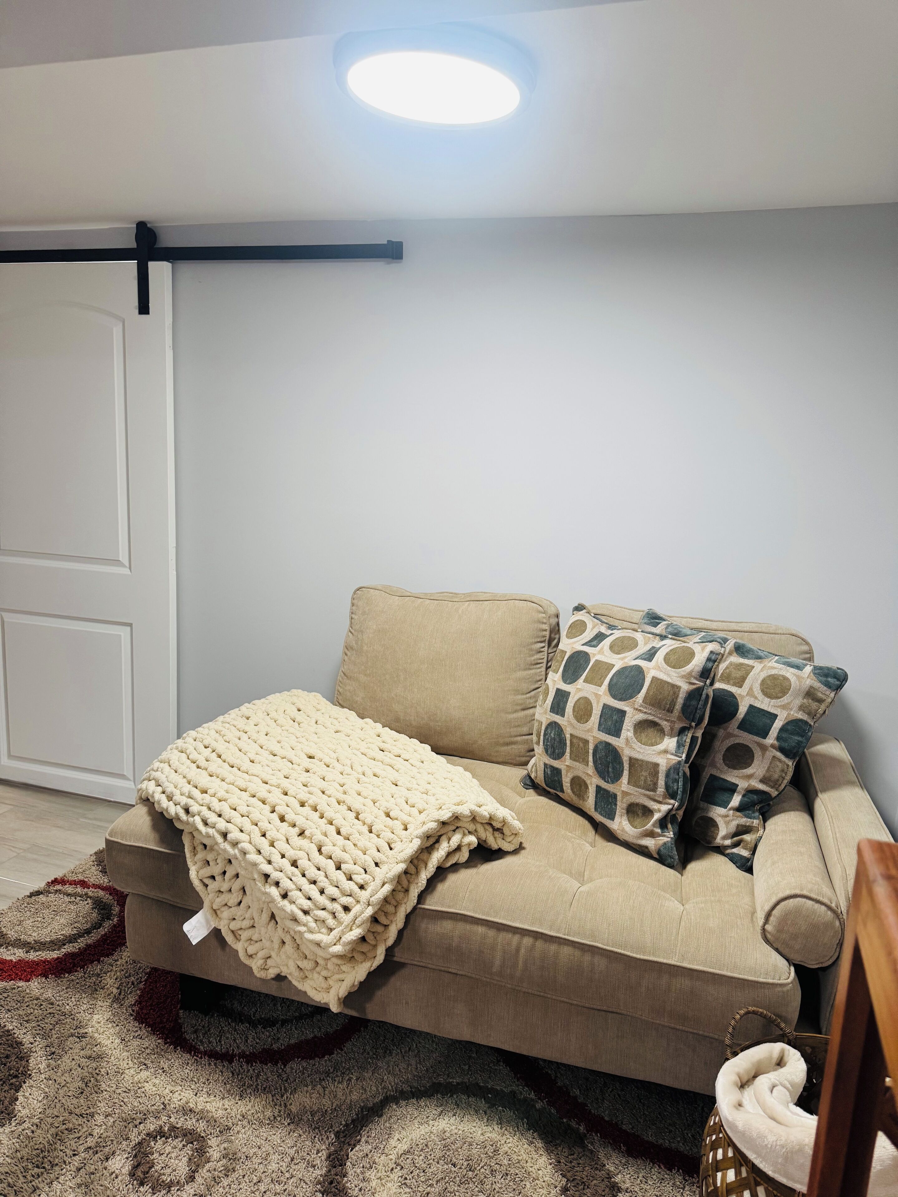 Un soggiorno con un divano beige, una coperta lavorata a maglia, cuscini decorati, pavimento in legno e una porta bianca.