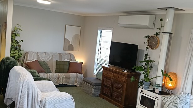 A cozy living room with a sofa, a dining table with a plant, a TV, and a cabinet. - Cosy Papamoa Beachside Getaway in Prime Location. (Papamoa)