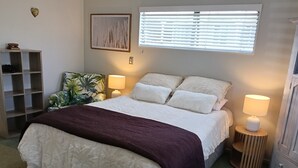 A neatly made bed with a white and purple bedspread, two bedside lamps, a window with blinds, a heart-shaped wall decoration, and a plant. - Cosy Papamoa Beachside Getaway in Prime Location. (Papamoa)