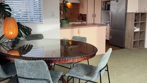 A modern kitchen with a central island, a dining area with a glass table, and grey upholstered chairs. - Cosy Papamoa Beachside Getaway in Prime Location. (Papamoa)