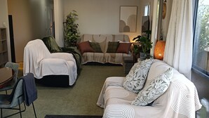 A modern living room with a sofa, a chair, and a bed. There is a window with curtains, a plant, and a lamp. - Cosy Papamoa Beachside Getaway in Prime Location. (Papamoa)