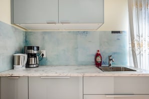 Una cocina con encimera de mármol, cafetera, hervidor, botella de detergente y pileta de acero inoxidable.