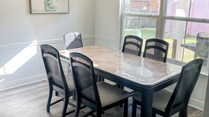 A dining area with a large table and six chairs, a hanging light fixture, and a window with a view of a backyard. - Charming 3-bedroom minutes from OWA (Foley)
