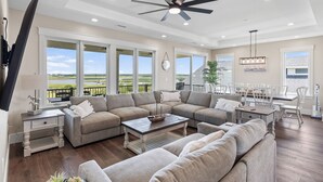 A spacious living room with a large sectional sofa, a dining area with white chairs, and a ceiling fan.