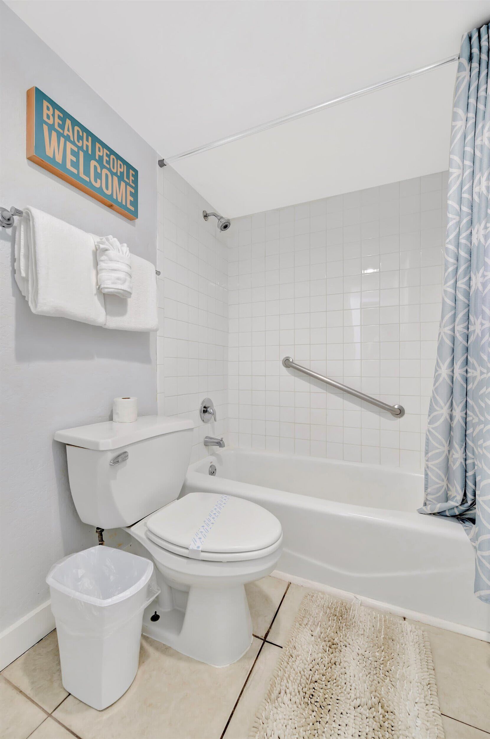 Une salle de bain équipée de toilettes, d’un bidet et d’une baignoire. On y trouve un porte-serviettes avec une serviette, une poubelle et un panneau indiquant : « LES AMANTS DE LA PLAGE SONT LES BIENVENUS ».