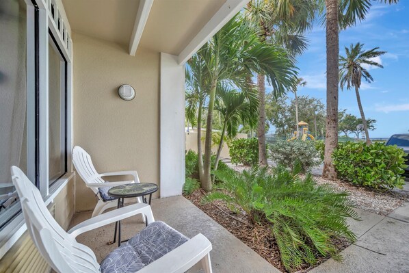 A balcony with white plastic chairs and a small table, overlooking a garden with palm trees and a yellow structure.