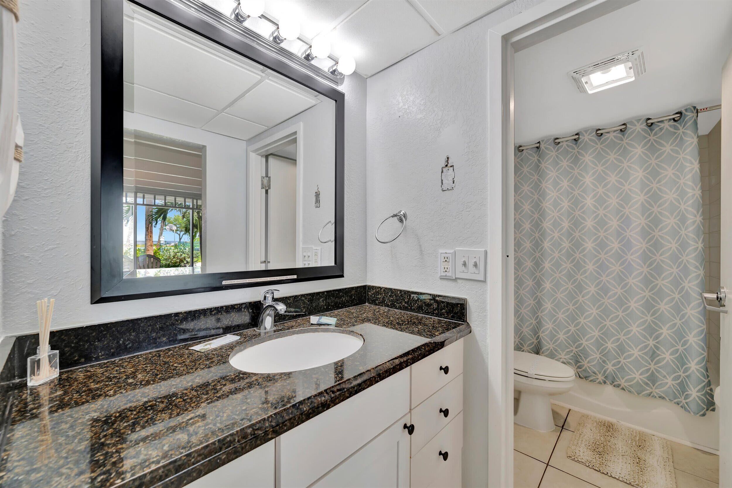 A bathroom with a granite countertop sink, a large mirror, a toilet, and a shower curtain.