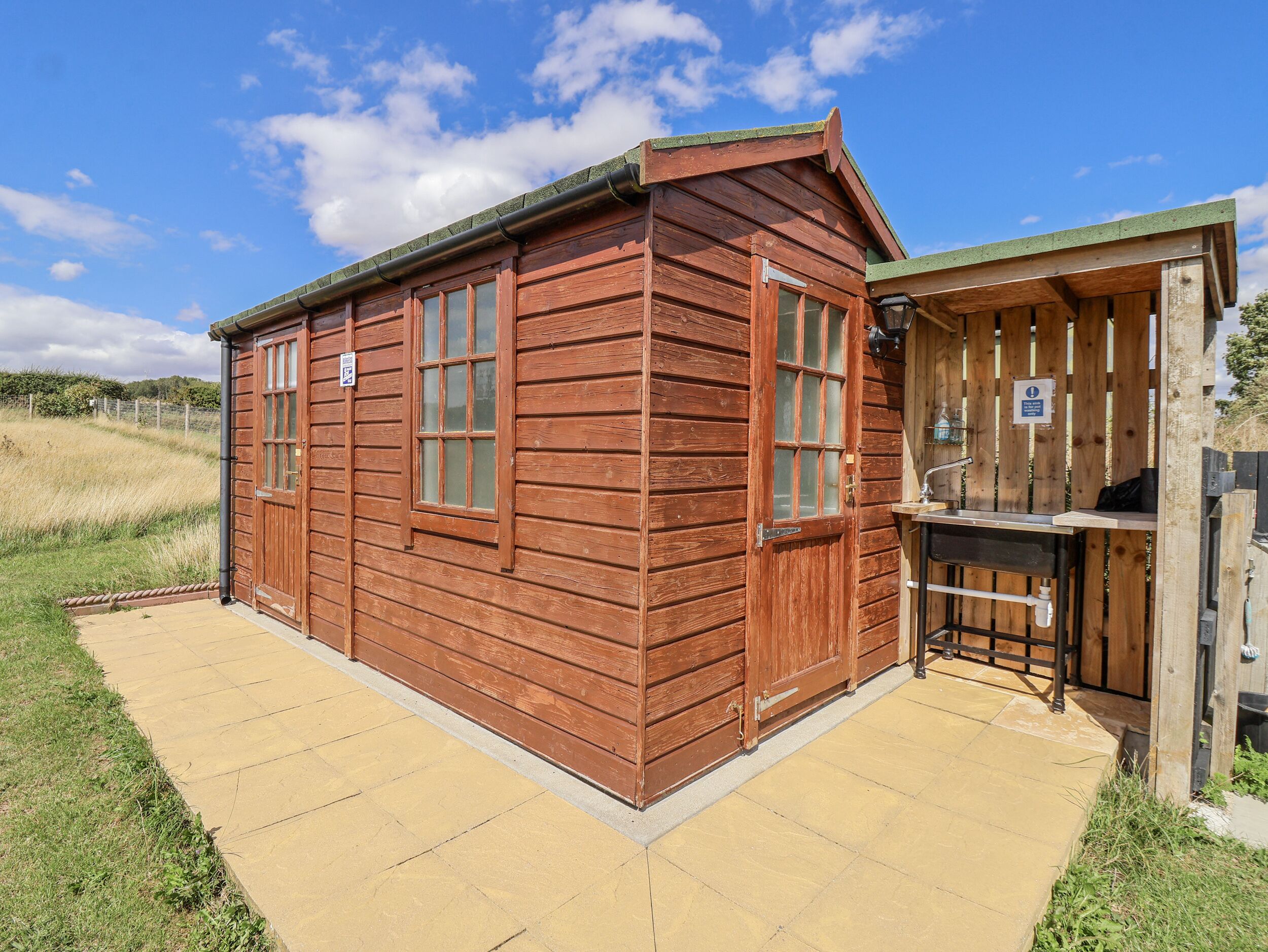 A wooden cabin with a large window and a door, a small outdoor sink, and a paved patio.