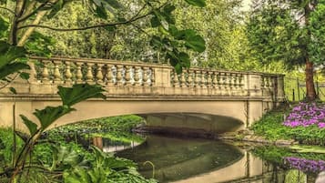 A stone bridge over a calm water body, surrounded by lush greenery and flowering plants.