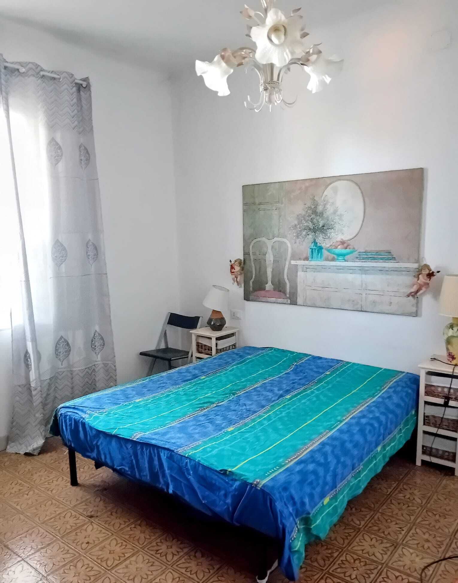 A bedroom with a bed, a desk, a chair, a painting, and a chandelier.