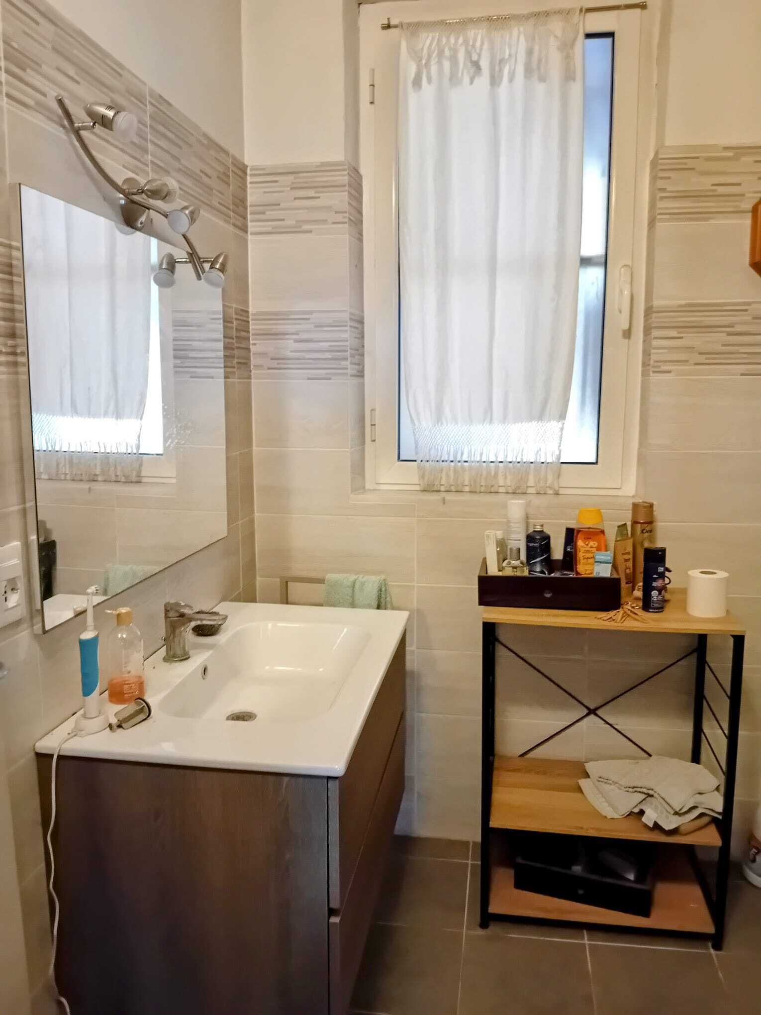 Une salle de bain dotée d’un grand miroir, d’une douche au design incurvé, d’une fenêtre avec un rideau blanc et d’une étagère en bois où sont rangés les articles de toilette.