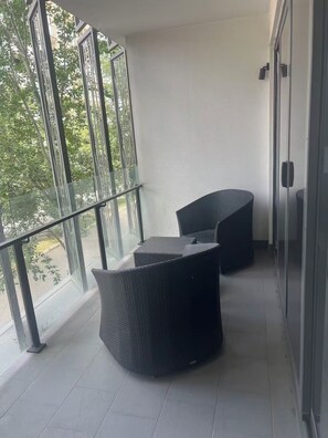 Un balcon avec deux fauteuils en osier noir, une balustrade en verre et une vue sur les arbres et les immeubles.
