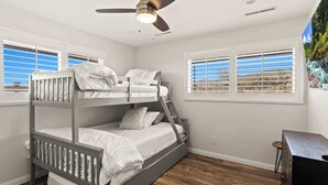 A bunk bed room with a ceiling fan, two windows with blinds, and a wooden floor.