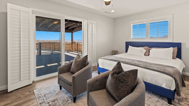 A bedroom with a large bed, two armchairs, a ceiling fan, and a view through a sliding door to an outdoor area.