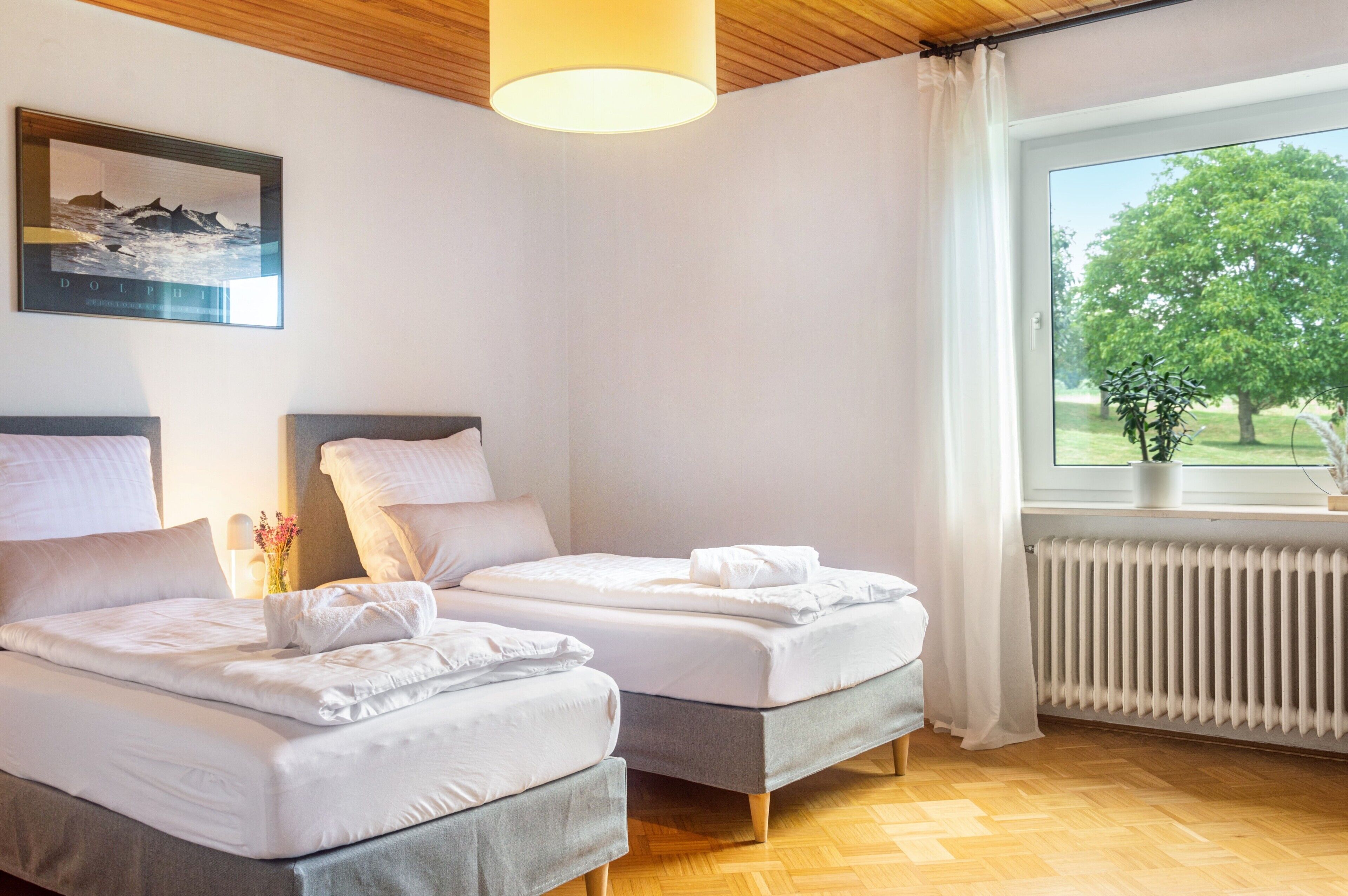 A hotel room with two beds, a window with a view of greenery, a radiator, and a framed picture on the wall.