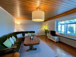 Een moderne woonkamer met een hoekbank, houten plafond, een eettafel, een televisie en een groot raam.