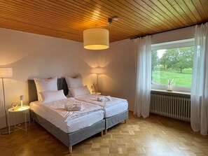 Een tweepersoonsbed met een grijs hoofdbord, nachtlampjes, een raam met uitzicht op groen en een houten plafond.